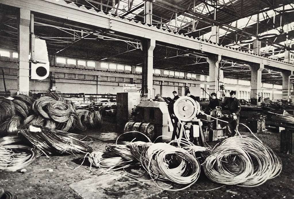 Hala produkcyjna z maszynami i pracownikami przy obróbce metali, stosy drutu w tle.