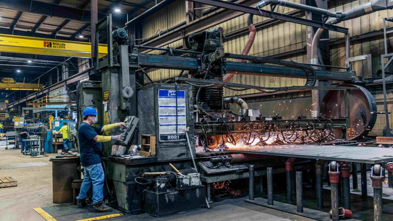 Pracownik w kasku ochronnym i rękawicach obsługujący maszynę przemysłową w dużej hali produkcyjnej, z widocznymi iskrami podczas obróbki metalu.