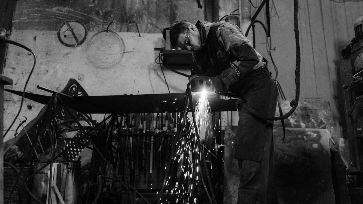 Spawacz w warsztacie przemysłowym wykonujący precyzyjne cięcie metalu, otoczony iskrami w surowym, industrialnym otoczeniu.