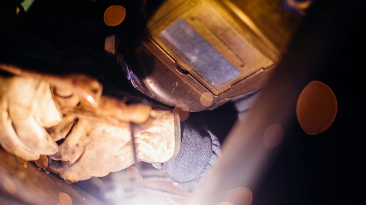Zbliżenie na spawacza podczas pracy – widoczne rękawice ochronne, przyłbica spawalnicza i jasne światło łuku spawalniczego.