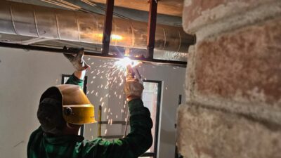 Spawacz spawa wspornik sufitowy przy kanale wentylacyjnym – widoczne iskry i drabina