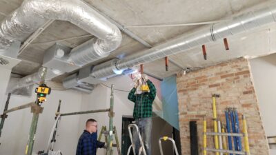 Dwóch spawaczy w pracy na drabinach i rusztowaniach w hali przemysłowej, montujących rurociągi wentylacyjne. Jeden spawa, drugi obserwuje. Obraz oddaje realia pracy w branży metalowej i instalacyjnej, podkreślając umiejętności, które wpływają na zarobki w 2026 roku.