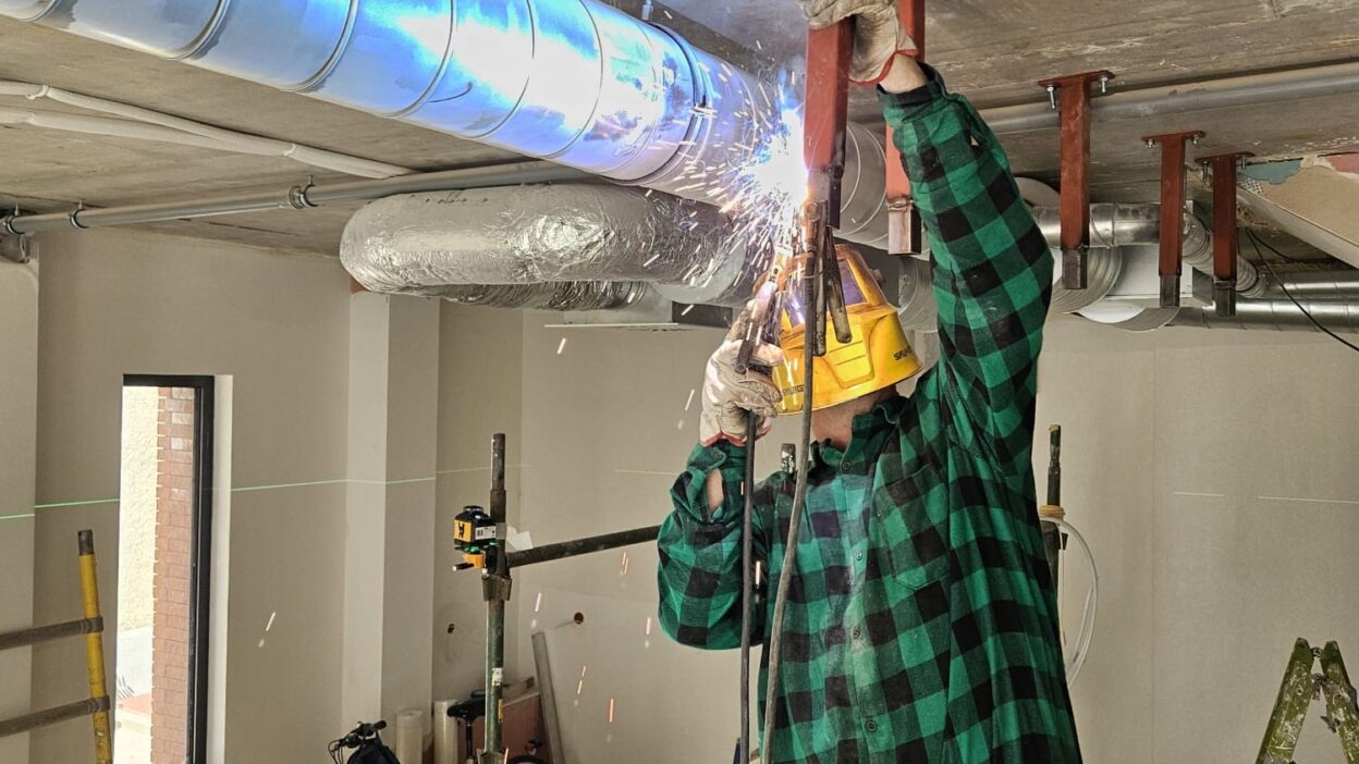 Pracownik w kasku ochronnym spawa metalowy wspornik do betonowego sufitu na placu budowy, wśród przewodów wentylacyjnych.