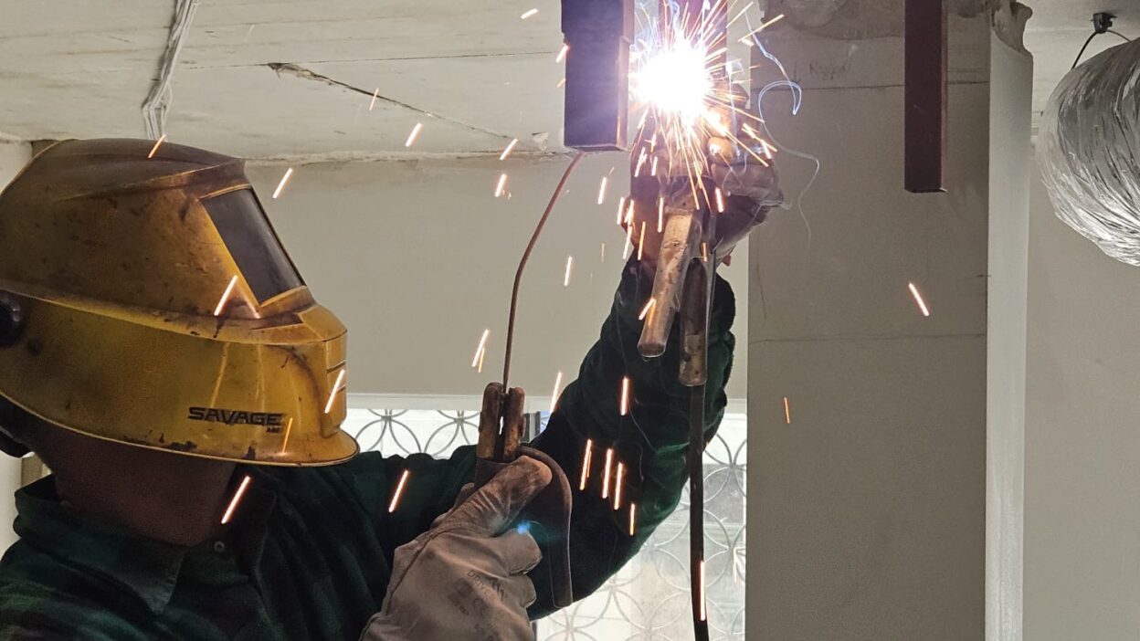 Zbliżenie na spawacza w żółtej masce ochronnej, który spawa metalową konstrukcję przymocowaną do betonowego sufitu, sypią się iskry.