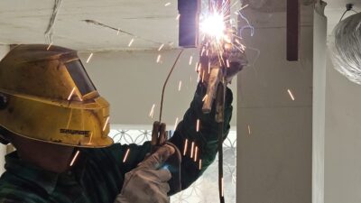 Zbliżenie na spawacza w żółtej masce ochronnej, który spawa metalową konstrukcję przymocowaną do betonowego sufitu, sypią się iskry.