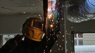 Spawacz w żółtej masce spawalniczej pracuje nad metalową konstrukcją wewnątrz budynku, sypią się snopy iskier.